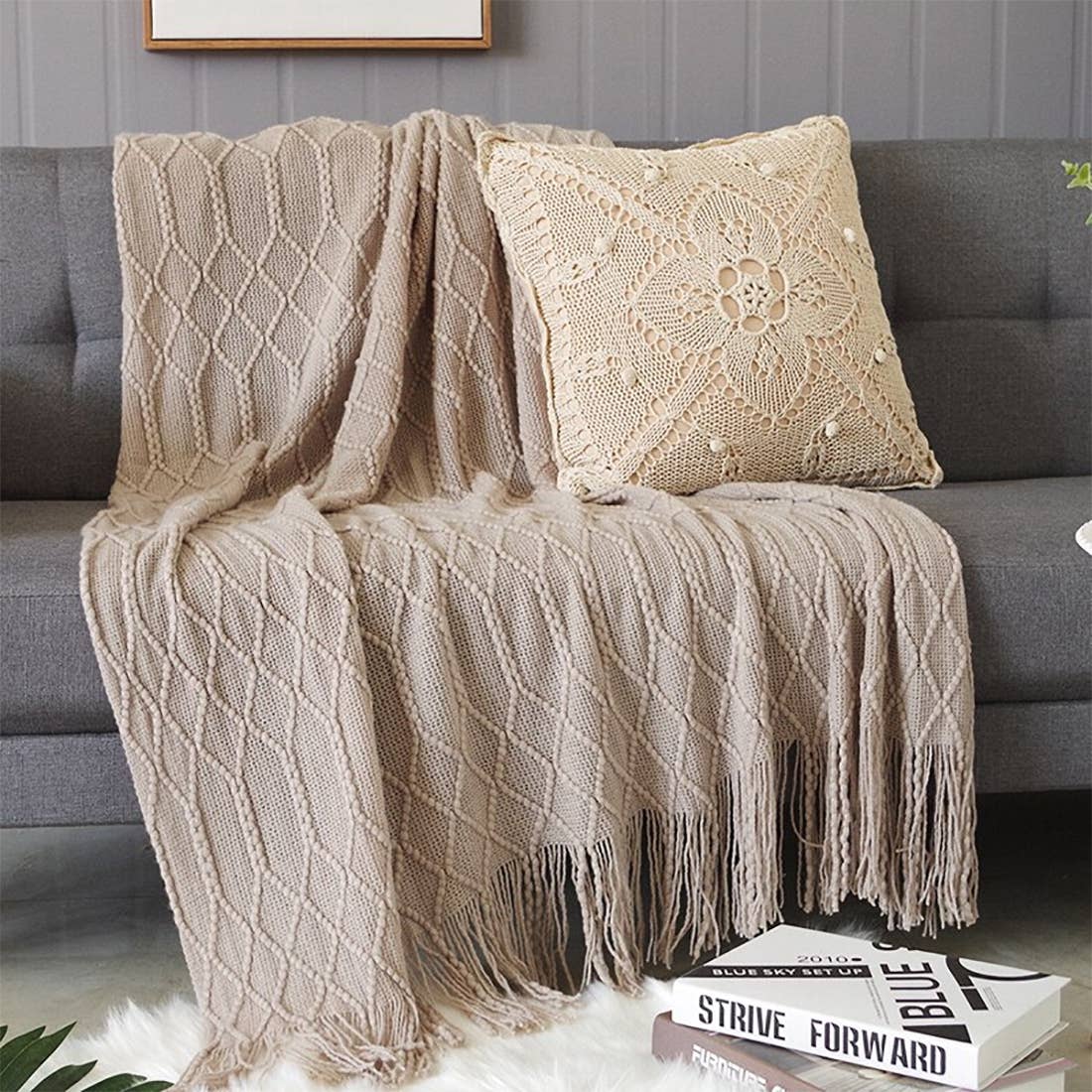 Beige knitted throw blanket and pillow on a gray sofa with books underneath.